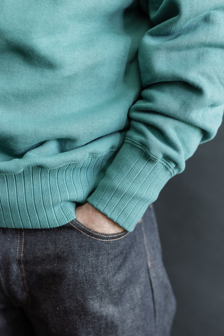 Close-up of male model’s right side, showing ribbed cuff and hem of blue-green sweatshirt.