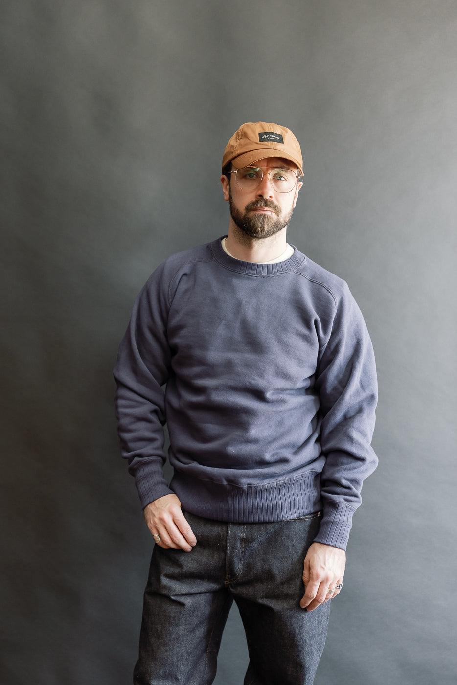 Bearded man in navy JG-CS24 Albert Sweat, inspired by Einstein's Princeton sweatshirt, showcases heritage style with freedom sleeves and circular knit, against a gray backdrop.
