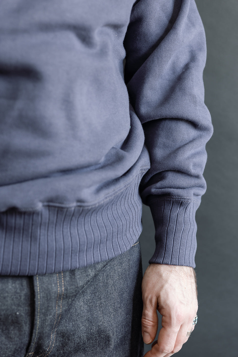 The lower half of a person wearing the JG-CS24 - The Albert Sweat in muted blue, showcasing the rib-knit hem and relaxed fit, paired with dark denim jeans.