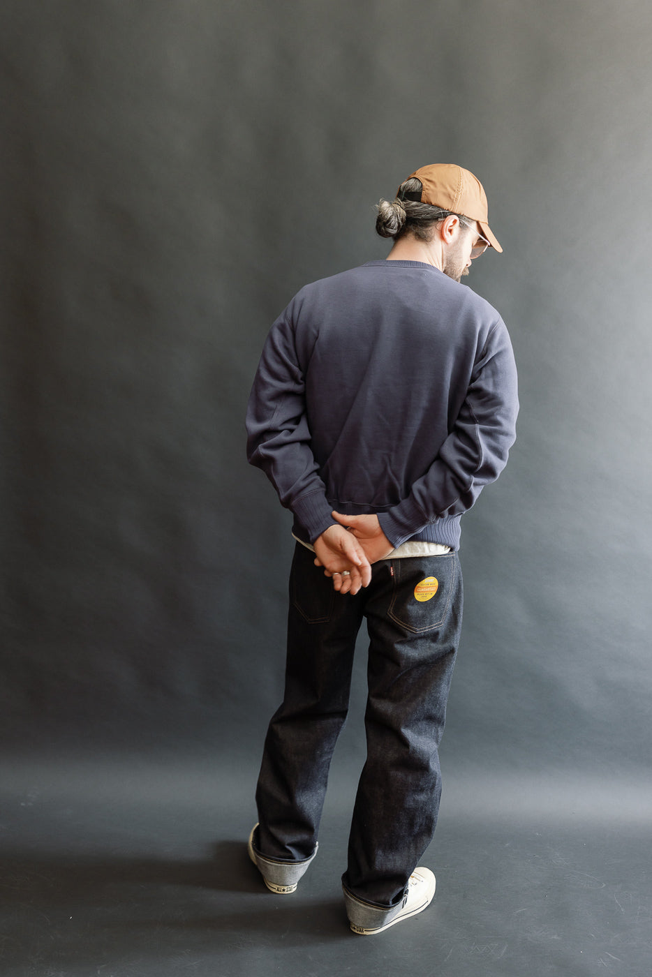 JG-CS24 - The Albert Sweat - Eggplant: Man in navy crewneck sweatshirt with freedom sleeve, relaxed posture, hands clasped behind, showcasing clean drape, paired with jeans and white sneakers.