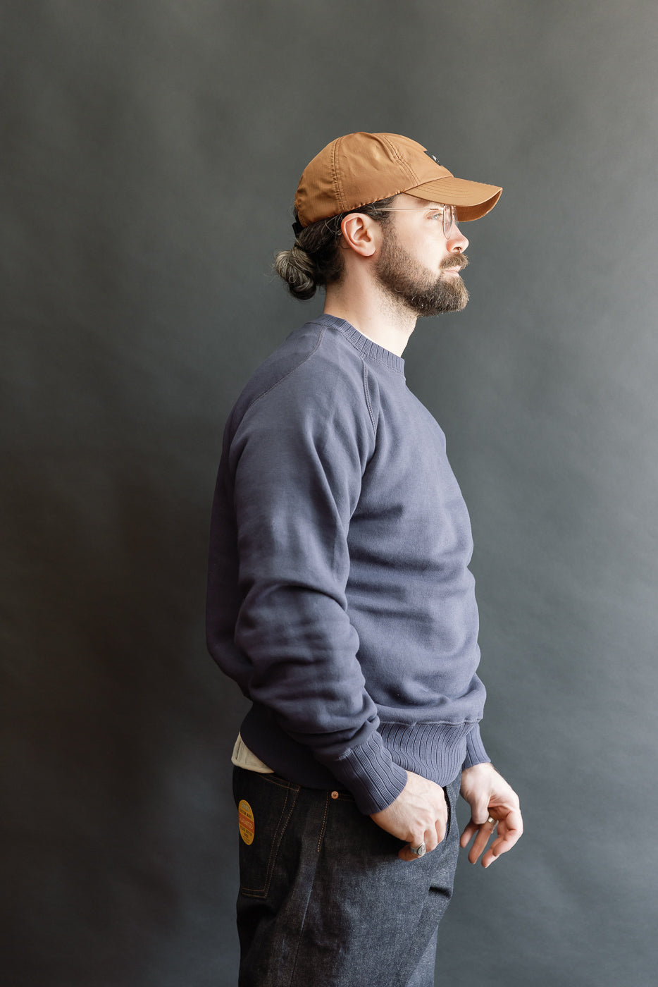 Bearded man in profile wearing JG-CS24 - The Albert Sweat - Eggplant, showcasing vintage-inspired navy blue crewneck sweatshirt with freedom sleeves, layered over a white T-shirt, paired with dark denim jeans.
