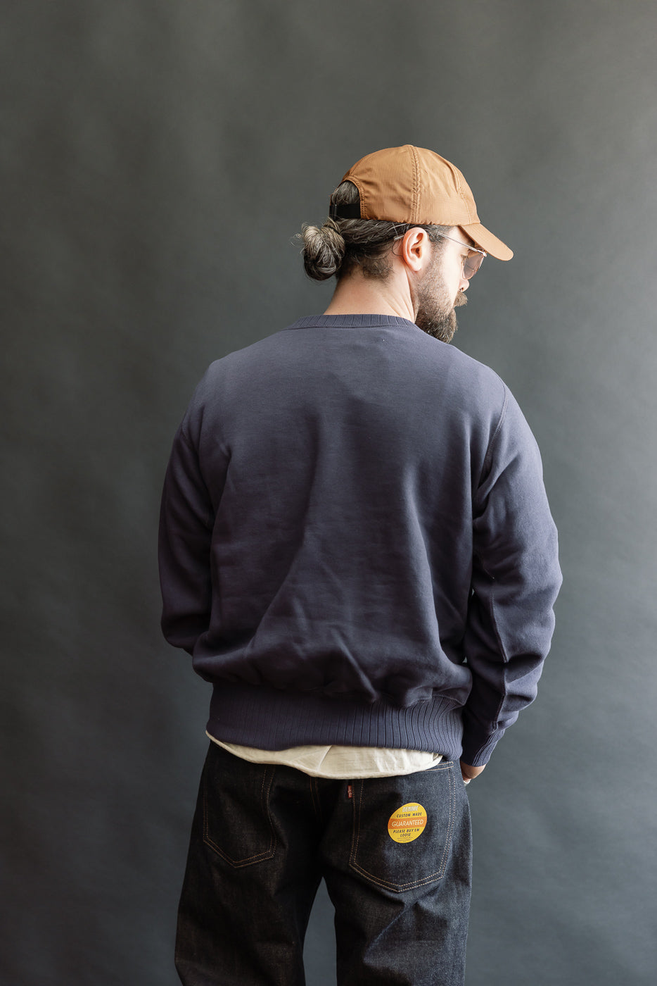 A casually-dressed man models the JG-CS24 Albert Sweat in a relaxed pose, showcasing its navy hue and classic crewneck fit, layered over a white T-shirt, with visible dark denim jeans.