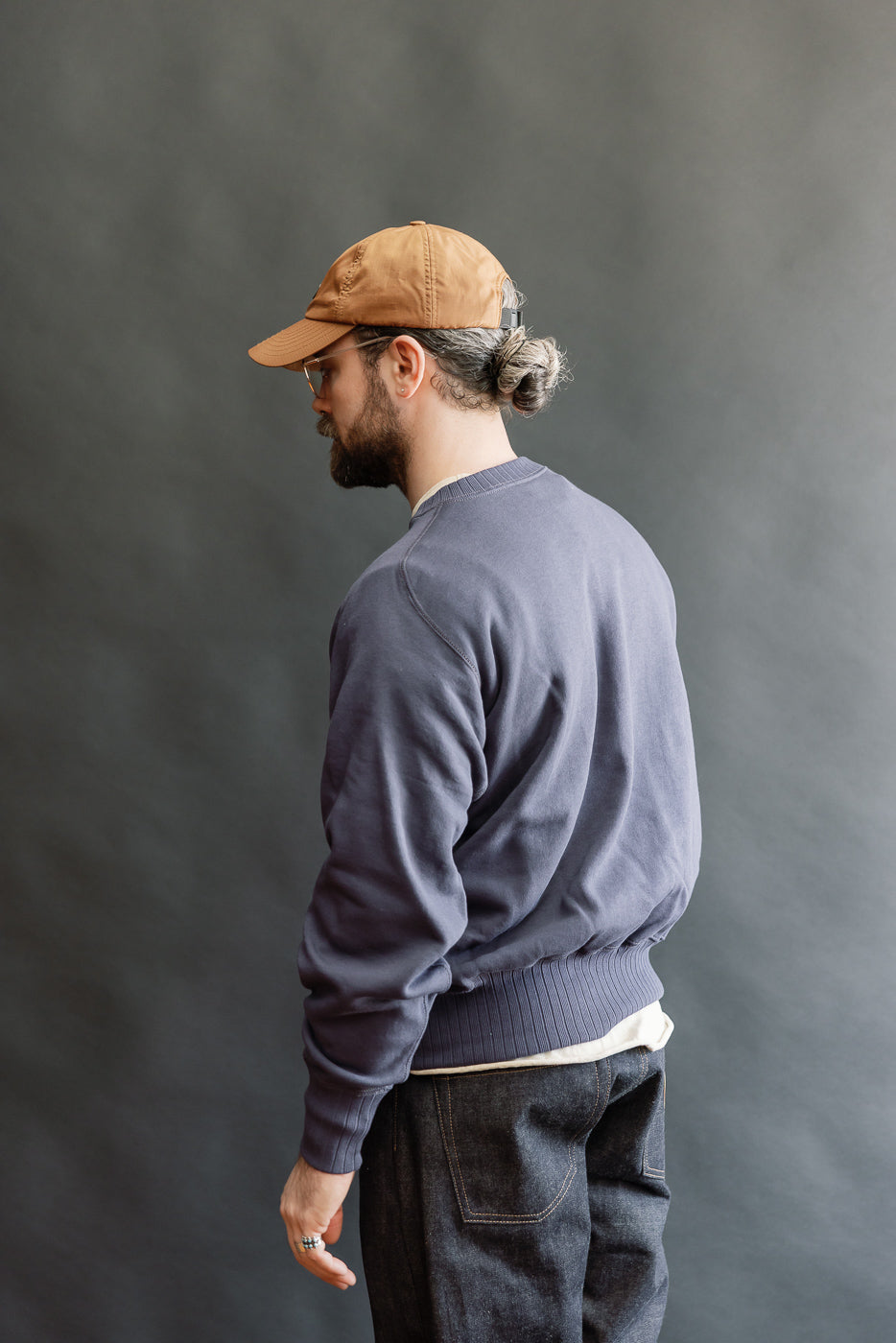 Man in profile wearing JG-CS24 - The Albert Sweat - Eggplant, a slate-blue crewneck sweatshirt with ribbed cuffs and hem, styled with dark denim jeans, showcasing a relaxed, vintage-inspired look.