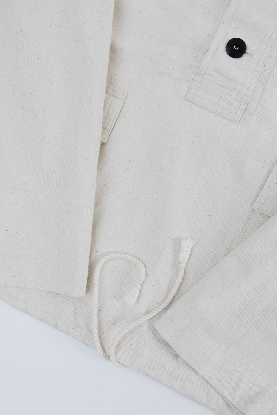 Close-up of off-white sailor's parka front hem, showing pocket, button, and drawstring detail.
