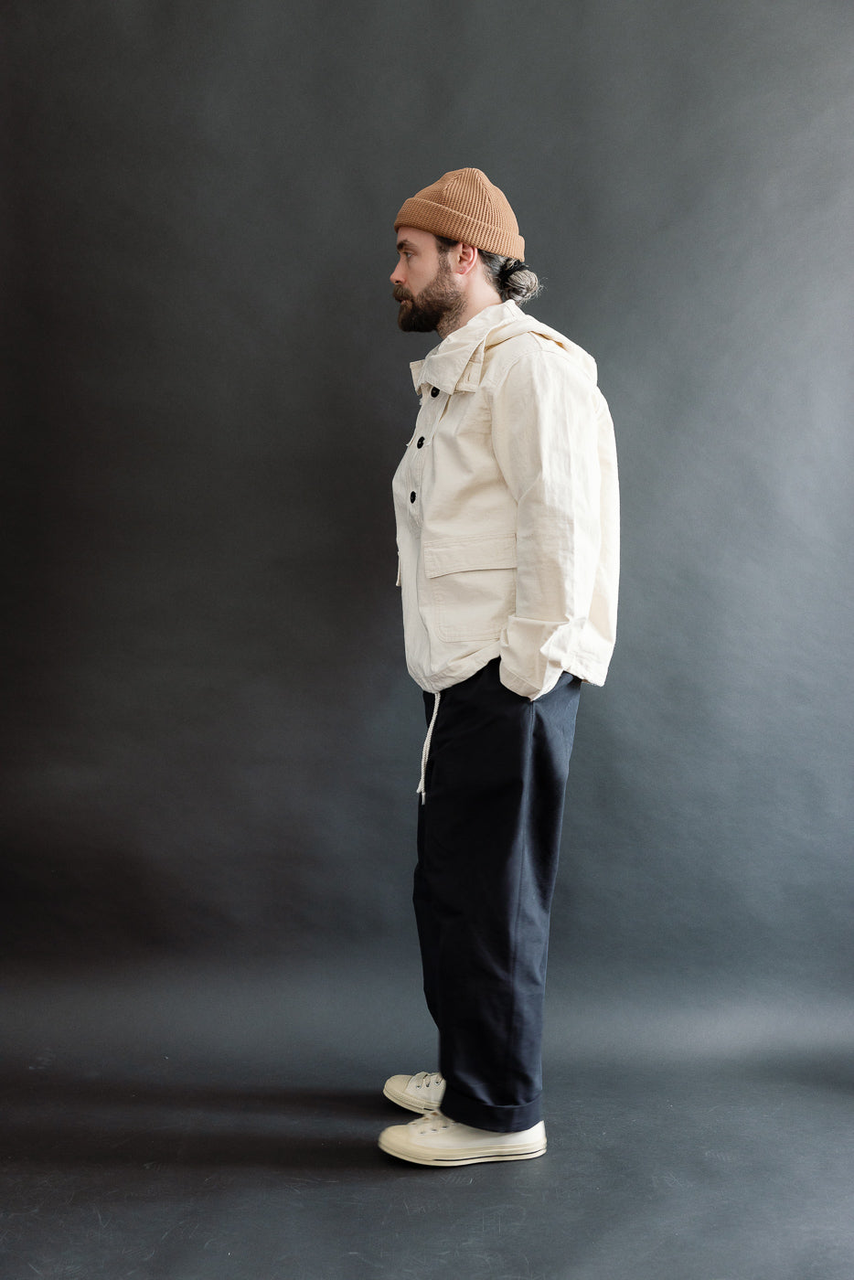 Full body side view of male model 5'9, 165lbs, athletic build wearing off-white sailor parka pullover, showcasing hood, front pockets, and button details.