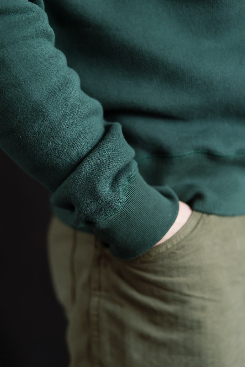 Close-up of male model's right side wearing Loop & Weft forest green crew neck, showing ribbed sleeve cuff and fabric texture. Model is 5'9