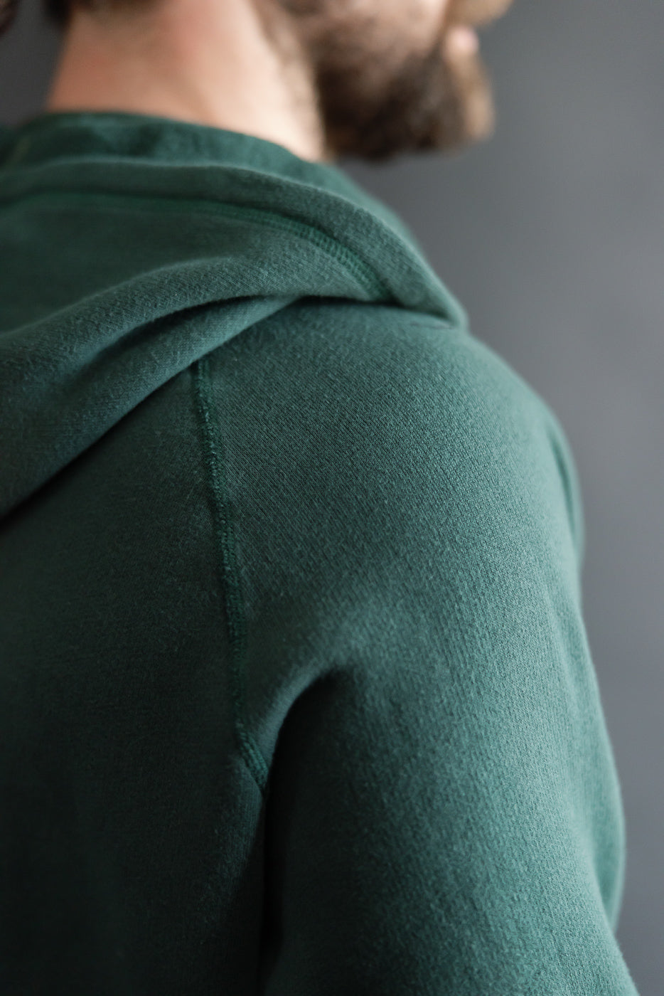 Close-up of male model's left shoulder wearing forest green raglan hoodie, showing fabric texture and hood seam.