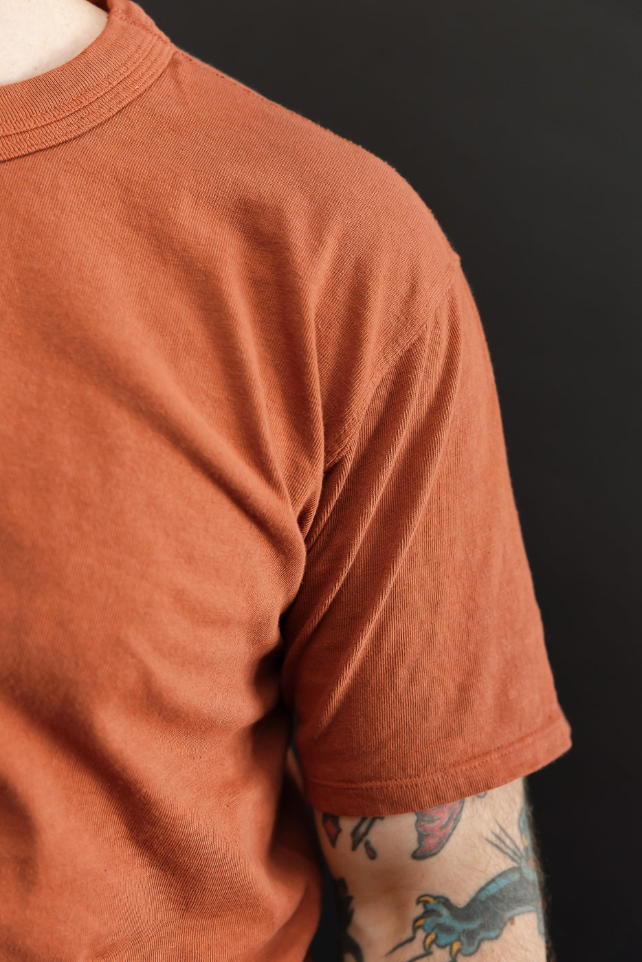 Close-up of male model's right shoulder, showcasing rust-colored tee with ribbed texture and sleeve hem.