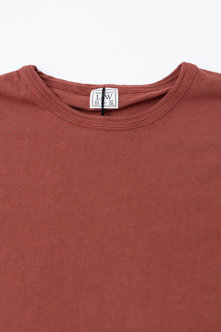 Close-up front view of rust-colored tee showing ribbed crew neck, fabric texture, and 'Made in Japan' tag detail
