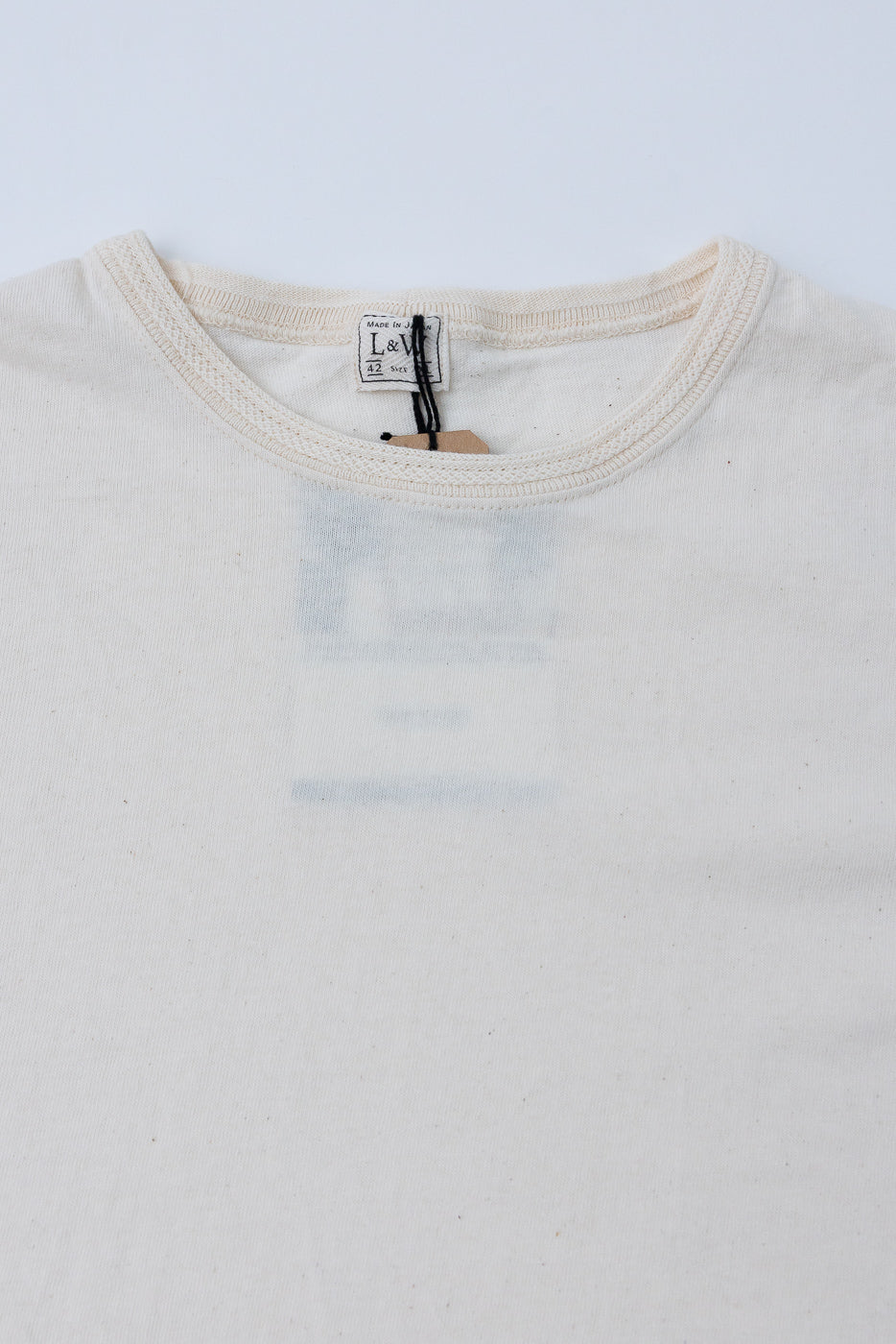 Close-up front view of cream crew neck tee, showing L&W tag, ribbed collar, and fabric texture