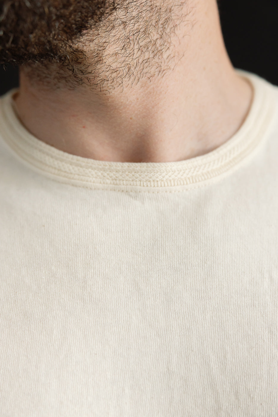 Close-up front view of male model's neck & crew neckline on off-white tee, showing ribbed collar detail