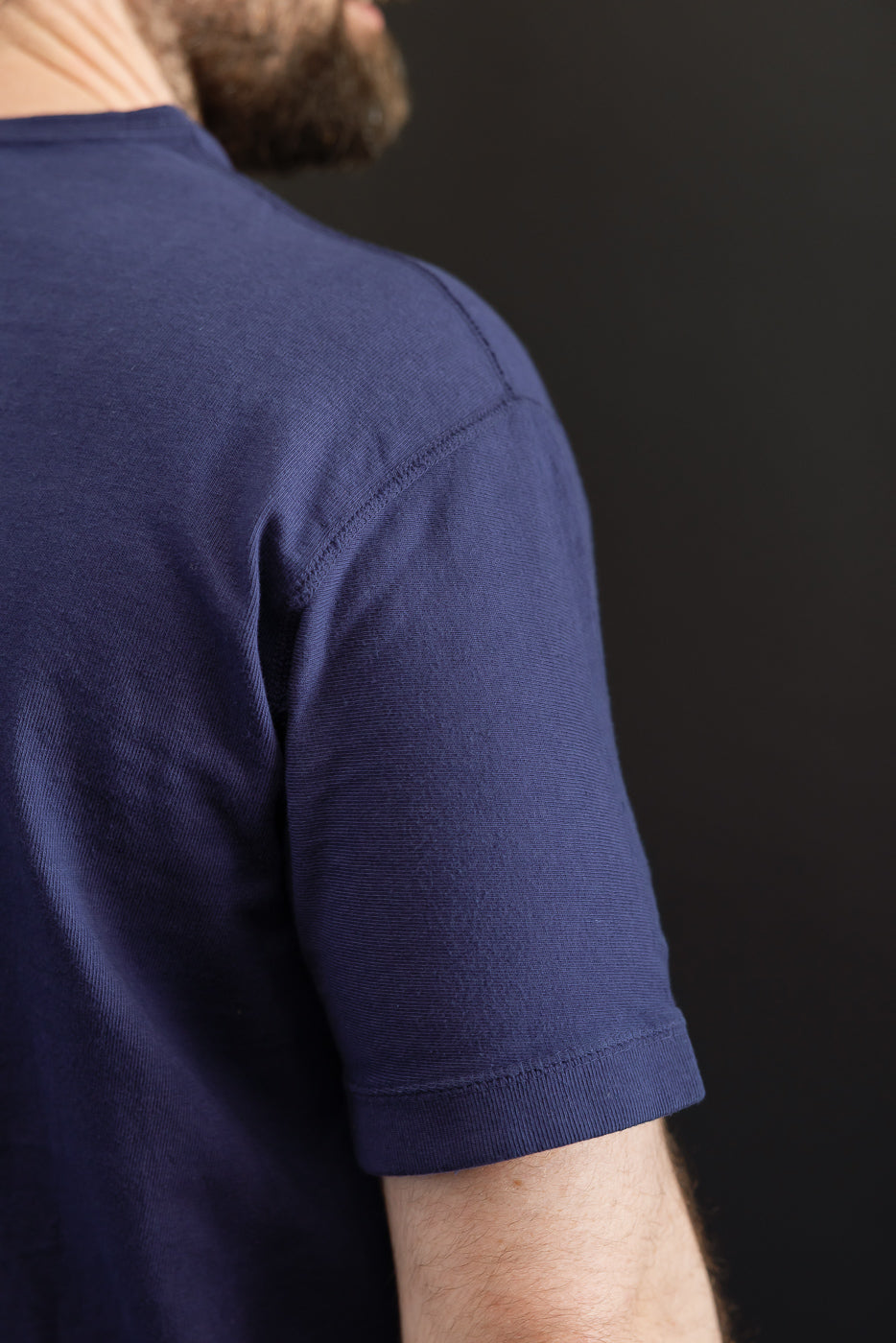Close-up side view of navy blue short sleeve tee, showing ribbed texture, sleeve hem, and shoulder seam on male model.
