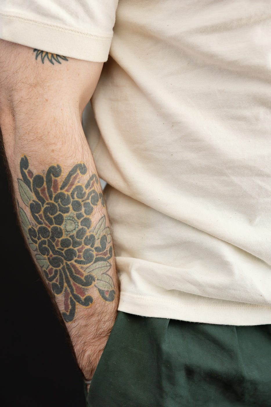 Close-up of male model's left arm showing off-shoulder t-shirt sleeve, tattoo detail, and fabric texture