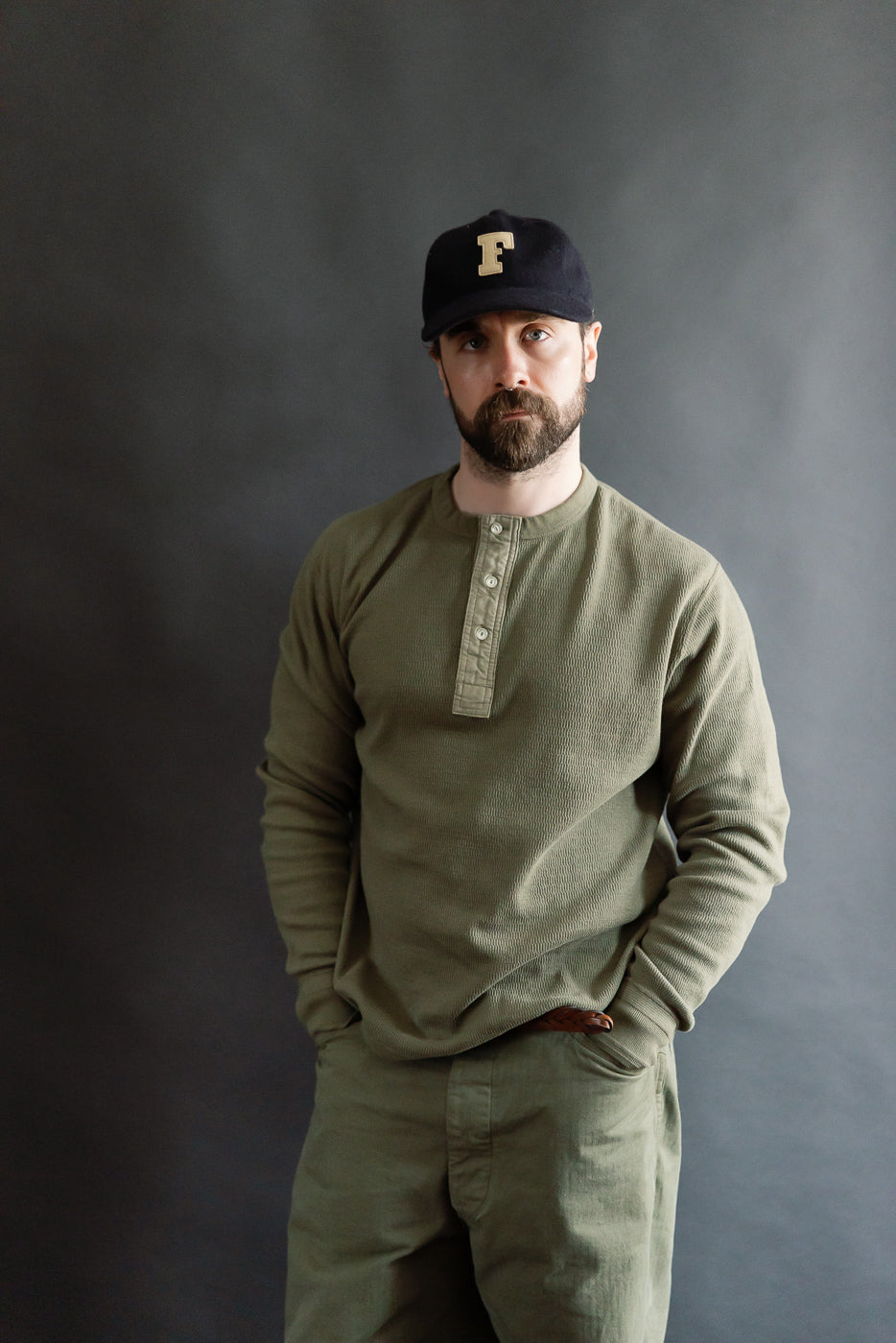 Front view mid-length of male model 5'9, 165lbs, athletic build wearing army olive Supima honeycomb knit long sleeve thermal Henley by Loop & Weft at Dant. Shows button placket, crew collar, textured fabric.