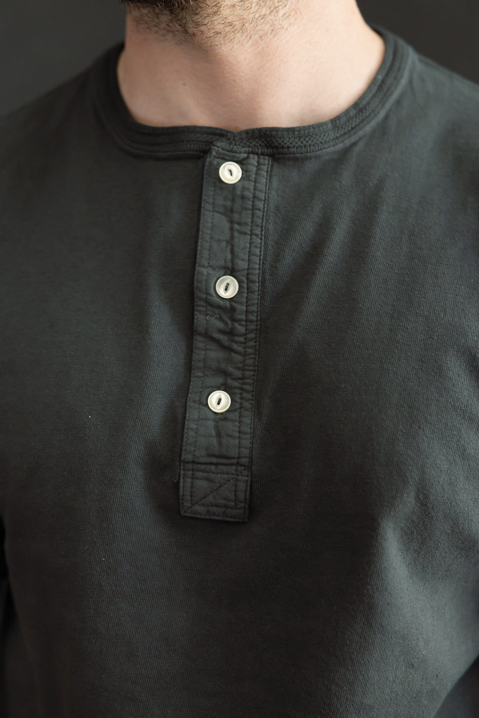Close-up front view of black henley shirt with white buttons, worn by male model, 5'9