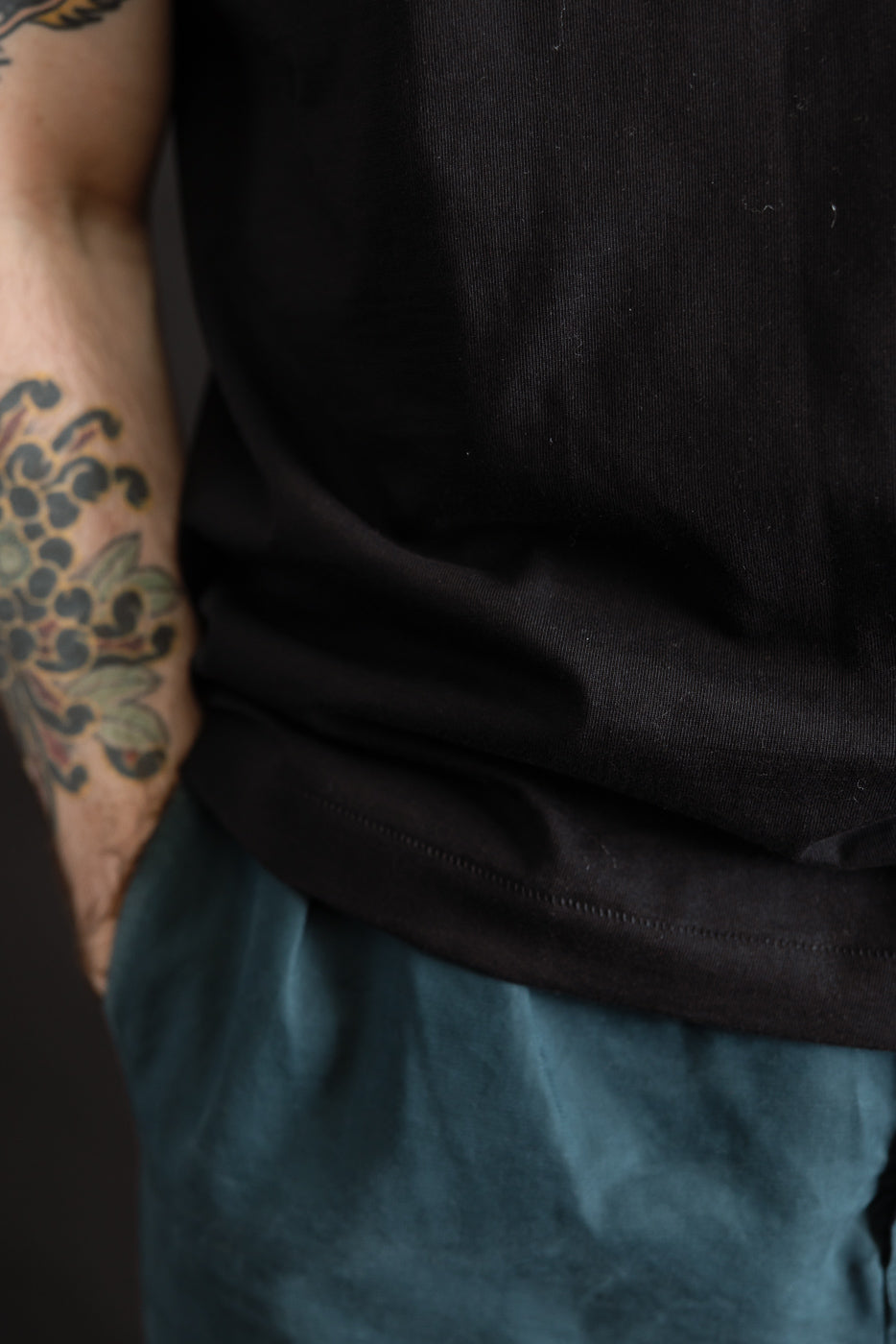 Close-up of a person wearing the 215.99 - 7.2oz Loopwheeled T-Shirt Classic Fit - Deep Black, showcasing its textured fabric and comfortable fit, with a hand casually placed in the pocket of teal pants.