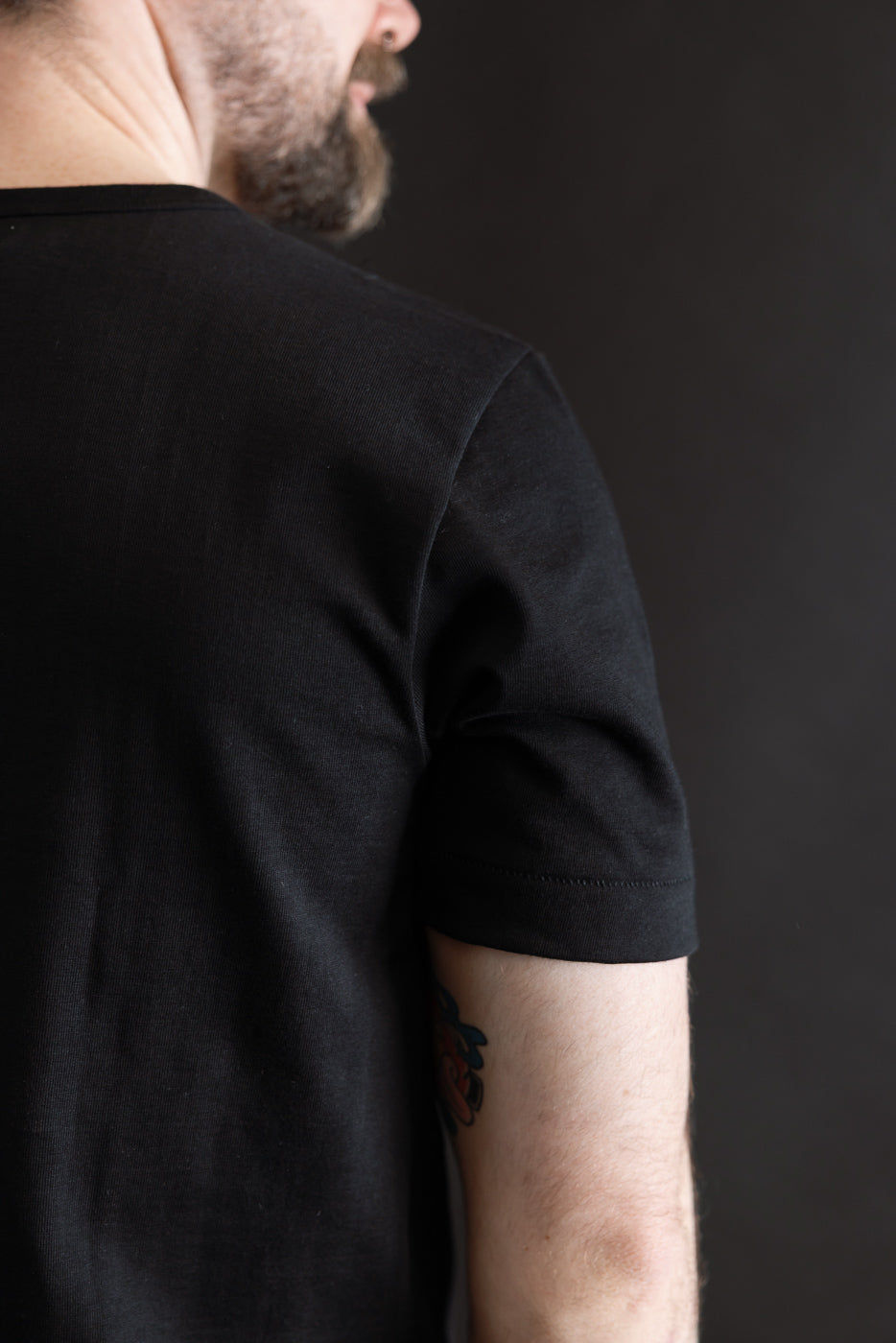 Person in profile wears the 215.99 - 7.2oz Loopwheeled T-Shirt Classic Fit - Deep Black. The tee's textured fabric and classic fit are highlighted, with a small tattoo visible on the upper arm.