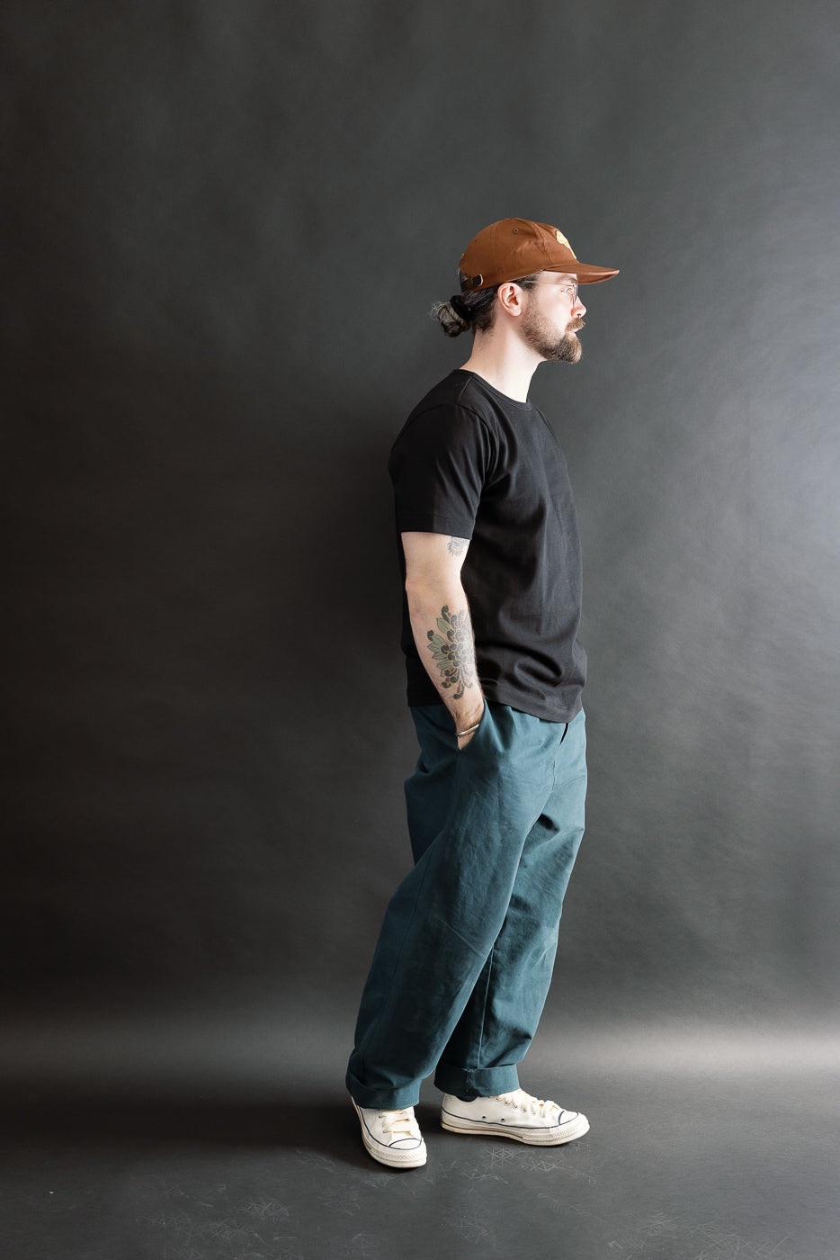 Full-length portrait of a man in profile wearing the 215.99 - 7.2oz Loopwheeled T-Shirt Classic Fit in Deep Black, showcasing its classic fit and subtle texture, paired with teal trousers and canvas sneakers.