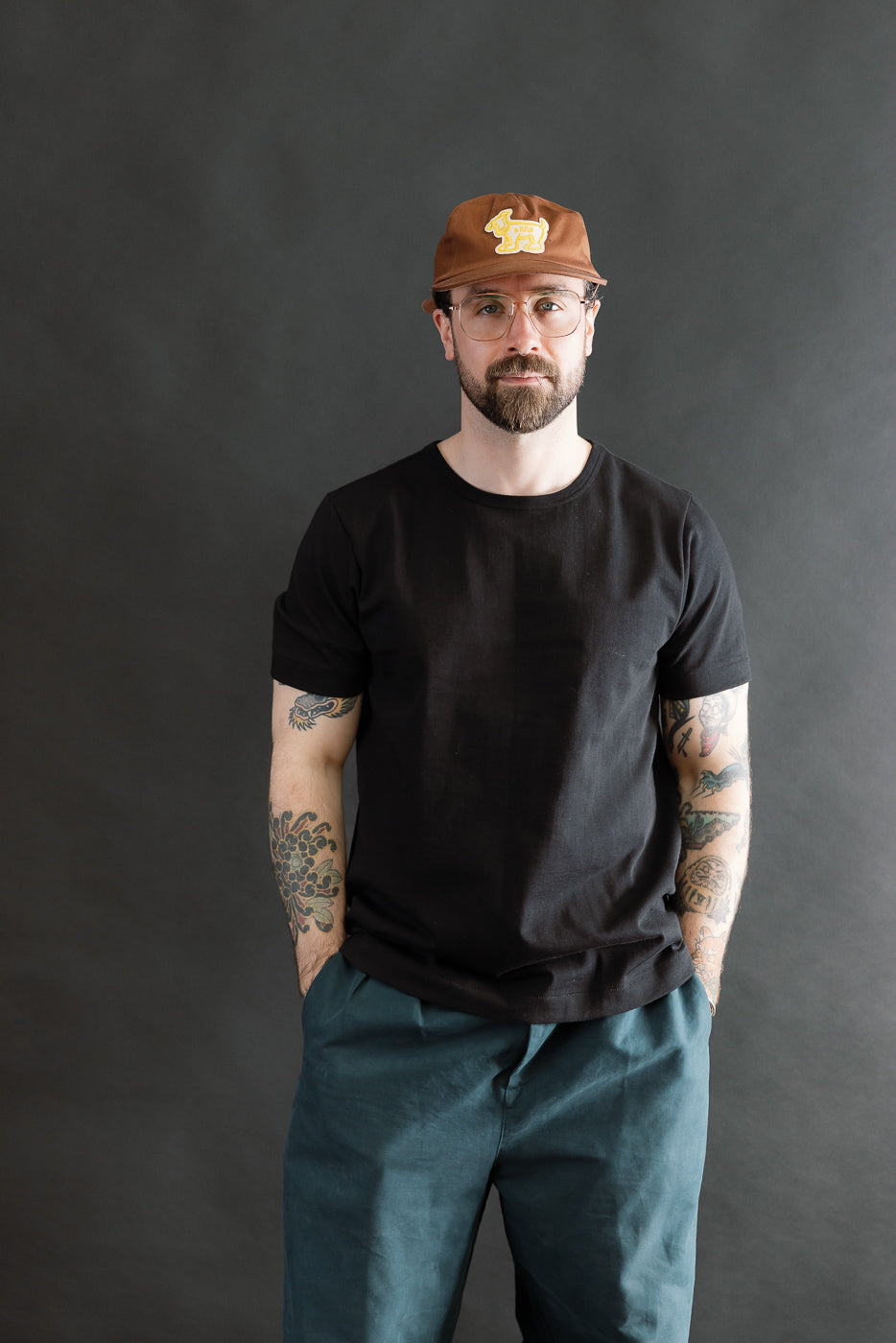 A man models the 215.99 - 7.2oz Loopwheeled T-Shirt Classic Fit - Deep Black, showcasing its subtle texture and comfortable fit, paired with teal pants and a brown cap, embodying Dant's premium menswear ethos.