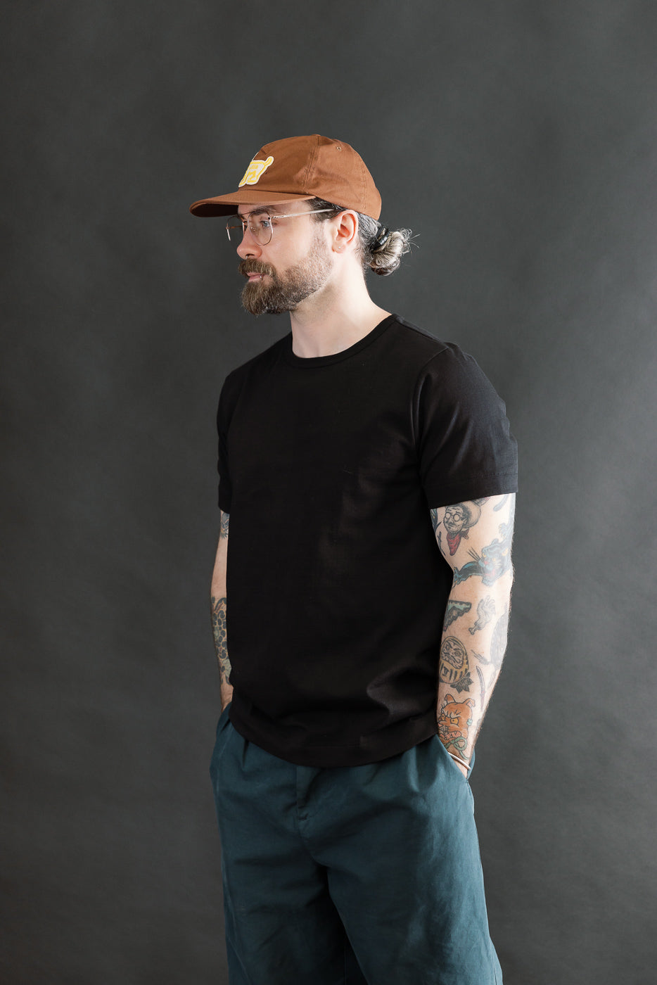 A model wearing the 215.99 - 7.2oz Loopwheeled T-Shirt Classic Fit in Deep Black, showcasing its classic fit and subtle loopwheel texture, styled with teal-blue pants and a brown baseball cap.