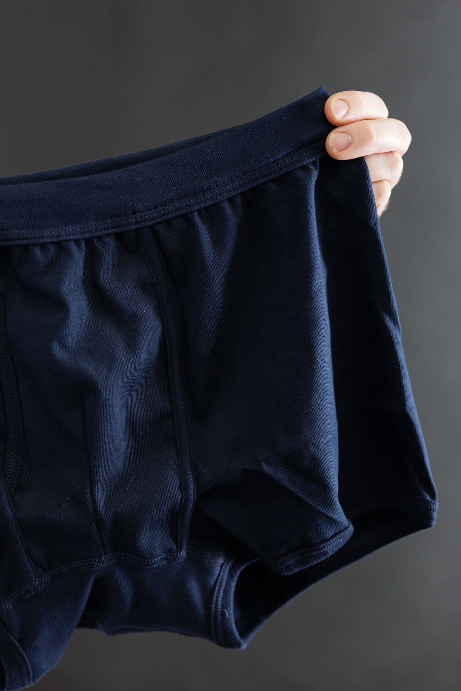 Hand holding the 254.66 - Loopwheeled Boxer Brief - Ink Blue, showcasing its elastic waistband and detailed stitching against a gray backdrop. The design emphasizes comfort and fit with a high thigh cut.