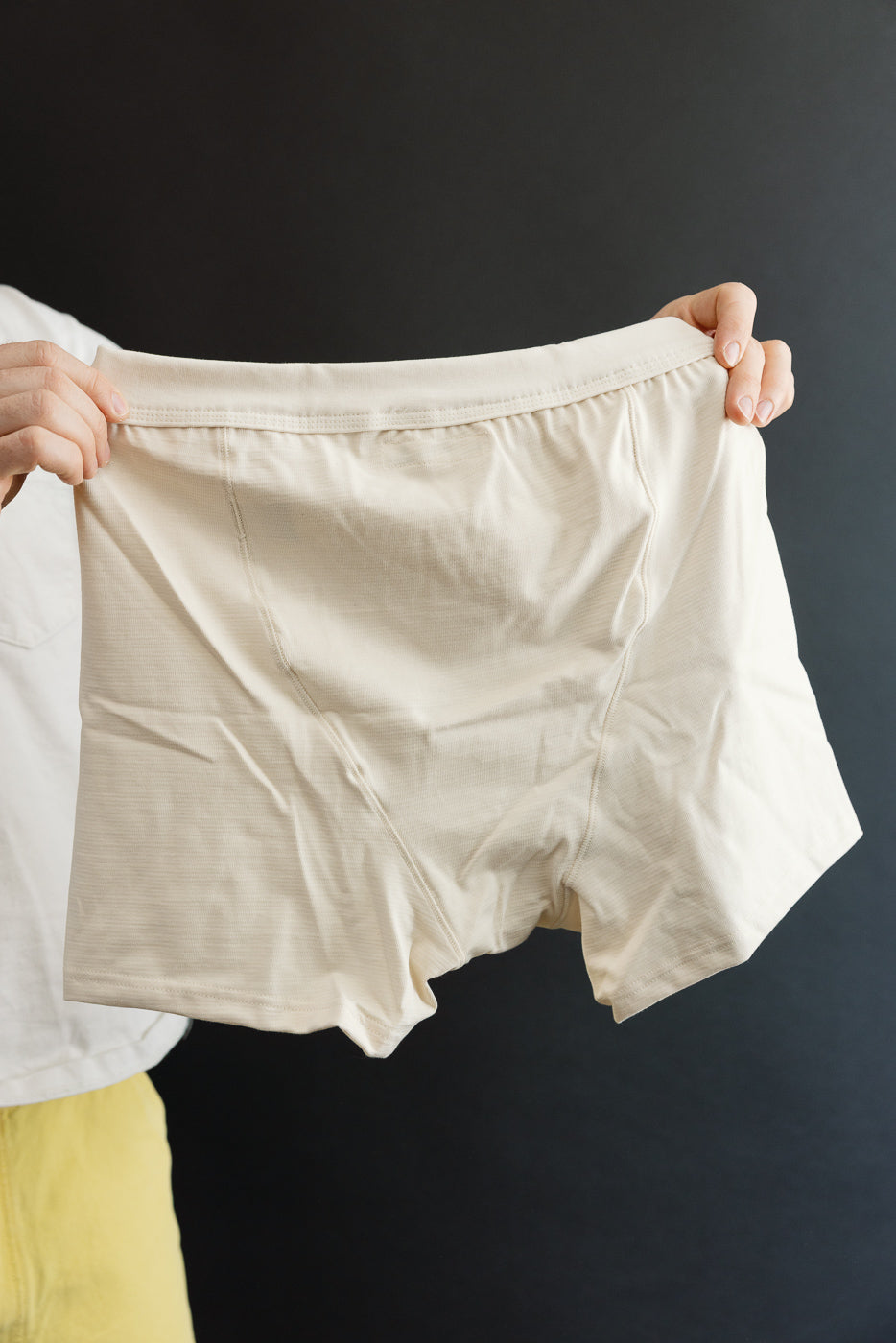 Close-up front view of natural color men's boxer shorts, fabric and waistband detail visible, held by a male model, 5'9