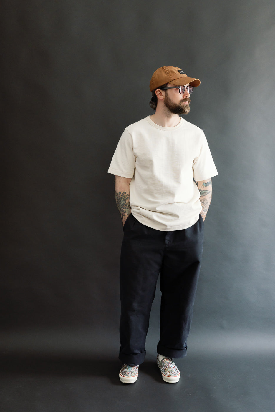 A bearded man models the 2S14.02 - 10.7oz Loopwheeled Heavy T-Shirt Relaxed Fit - Nature, showcasing its relaxed fit and detailed neckline, paired with black trousers and patterned slip-ons.
