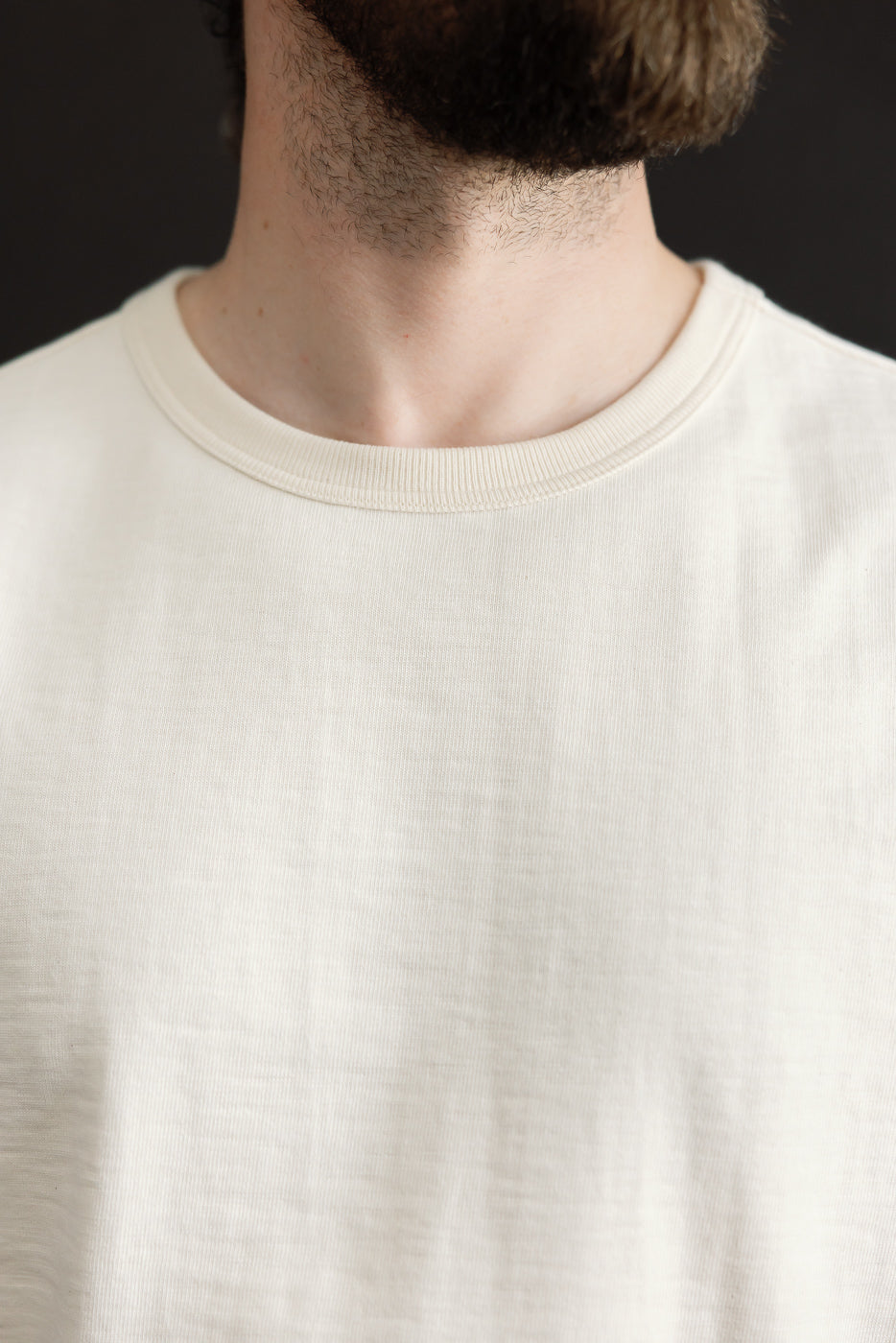 Close-up of an adult male wearing the 2S14.02 - 10.7oz Loopwheeled Heavy T-Shirt Relaxed Fit - Nature, showcasing its detailed neckline and slight slub structure against a dark background.