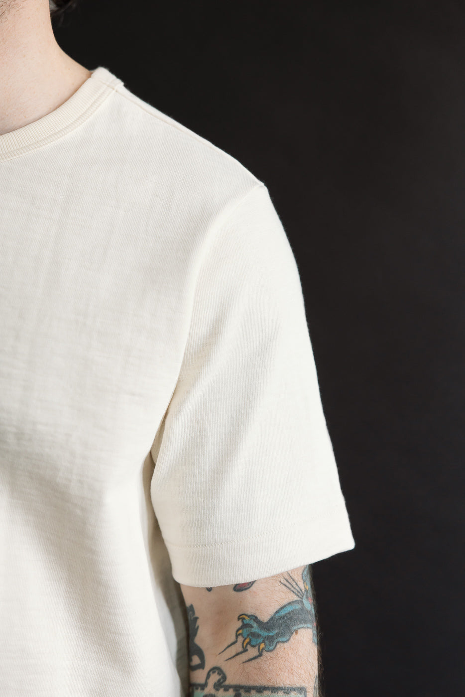 The 2S14.02 - 10.7oz Loopwheeled Heavy T-Shirt Relaxed Fit - Nature, featuring a slub texture, is showcased partially on a tattooed upper arm, highlighting its soft, premium craftsmanship.