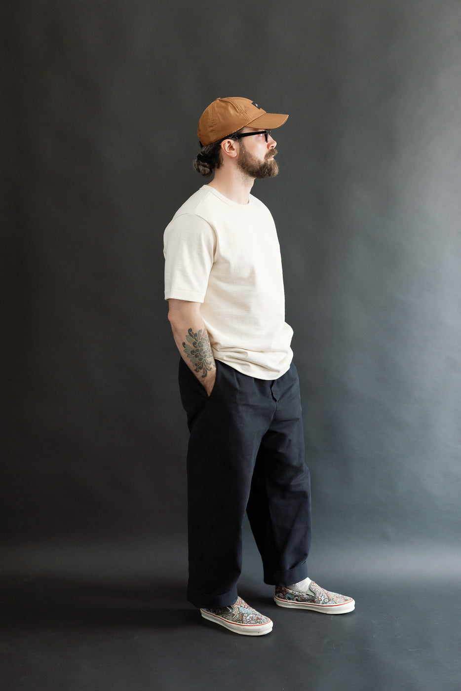 Man in profile wearing the 2S14.02 - 10.7oz Loopwheeled Heavy T-Shirt Relaxed Fit - Nature, tucked into dark trousers, showcasing a floral tattoo. His casual look is completed with patterned slip-on shoes.