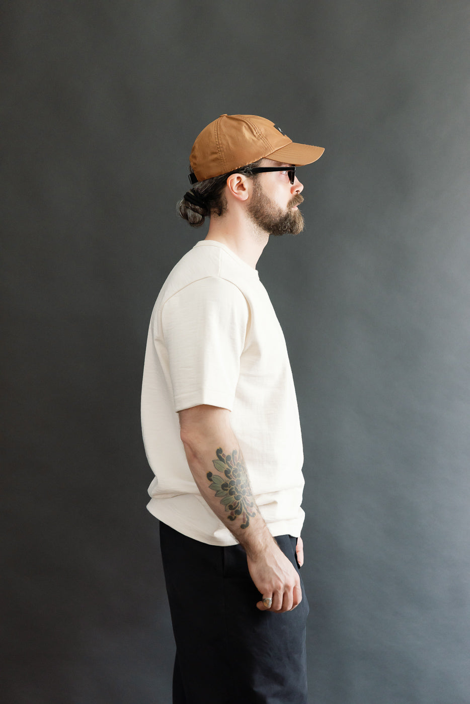 A bearded man models the 2S14.02 - 10.7oz Loopwheeled Heavy T-Shirt Relaxed Fit - Nature, showcasing its relaxed fit, detailed neckline, and slightly slubbed texture against a dark-gray backdrop.