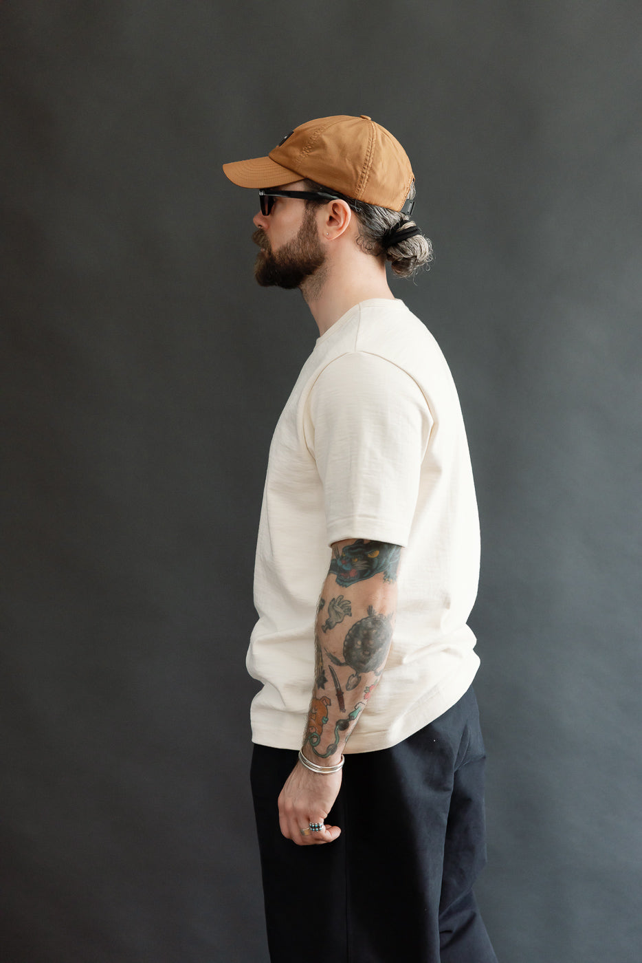 Man in profile wearing the 2S14.02 - 10.7oz Loopwheeled Heavy T-Shirt Relaxed Fit - Nature, showcasing its relaxed fit and detailed neckline. His tattooed arm adds visual contrast to this minimalist look.