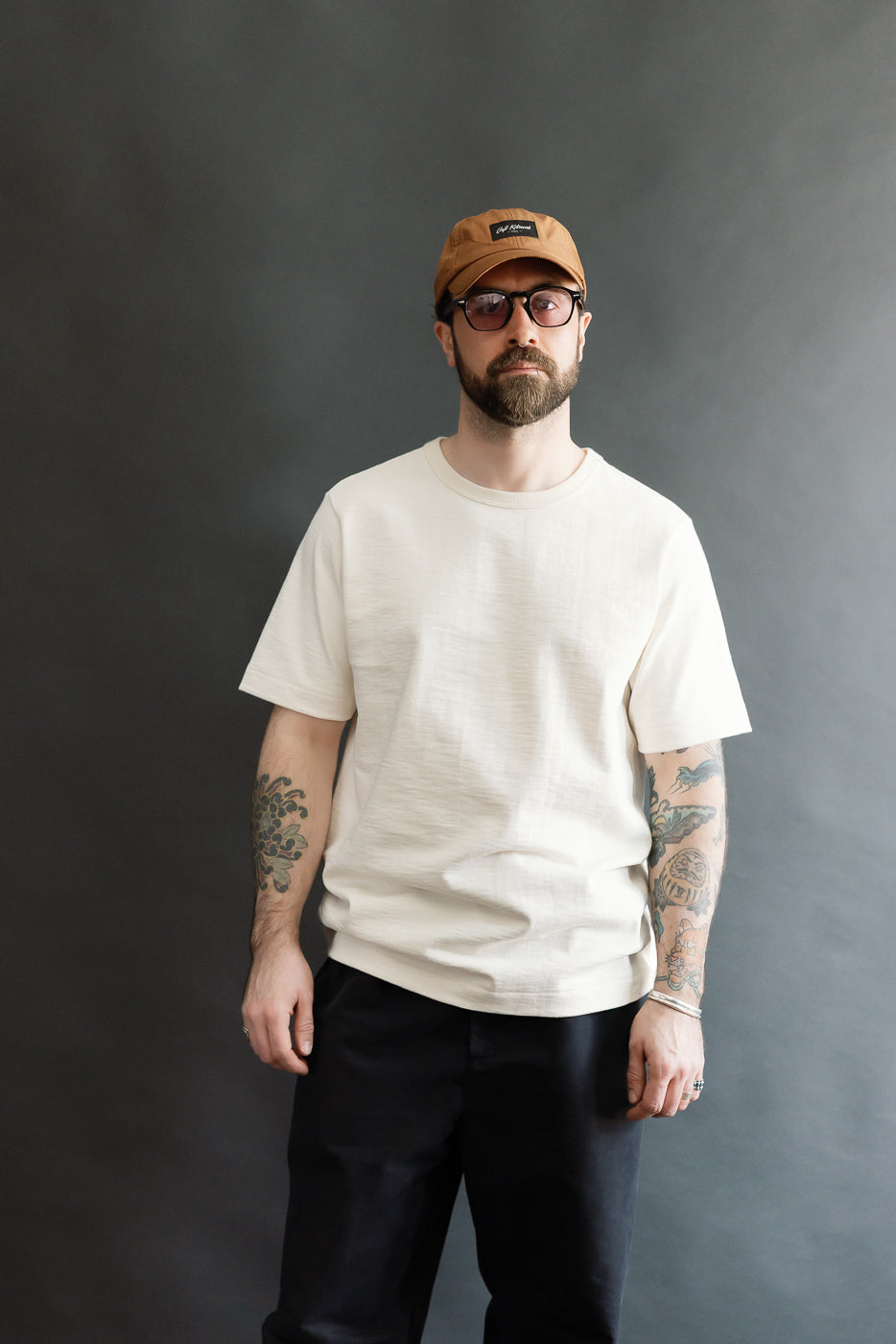A man models the 2S14.02 - 10.7oz Loopwheeled Heavy T-Shirt Relaxed Fit - Nature, featuring a light-beige, textured finish, highlighting its casual, premium design. He pairs it with black pants and accessories.