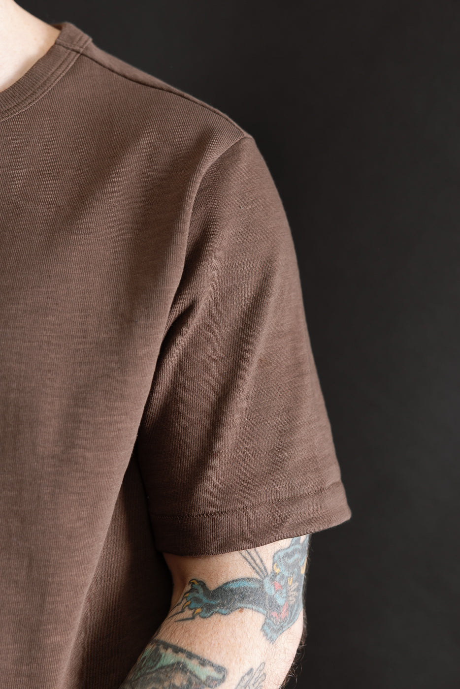 Close-up of male model's right front shoulder and sleeve on chocolate heavy T-shirt, showing fabric texture. Model is 5'9