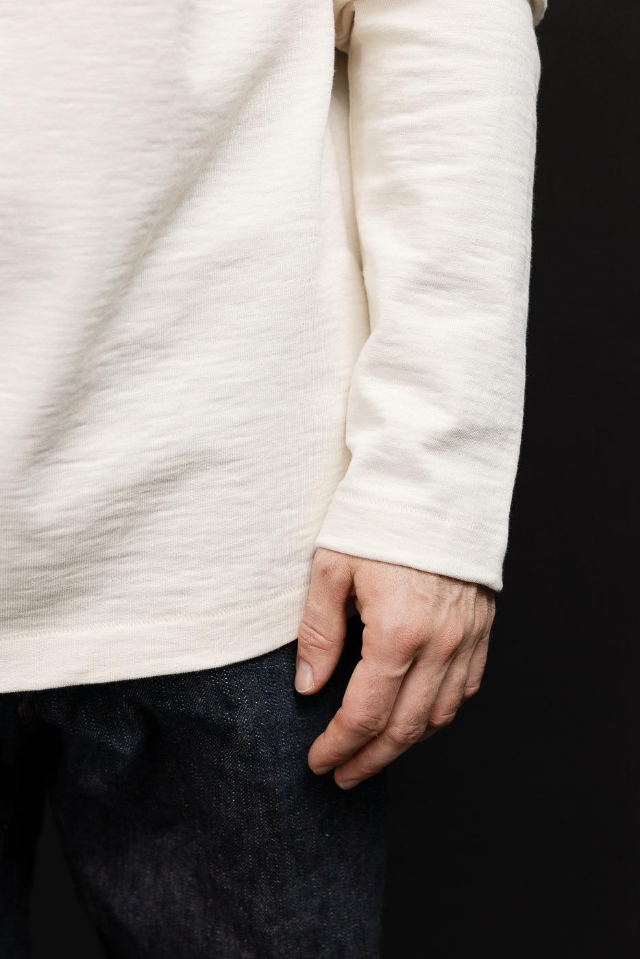 Close-up side view of a male model's cream long-sleeve shirt and dark denim pants.