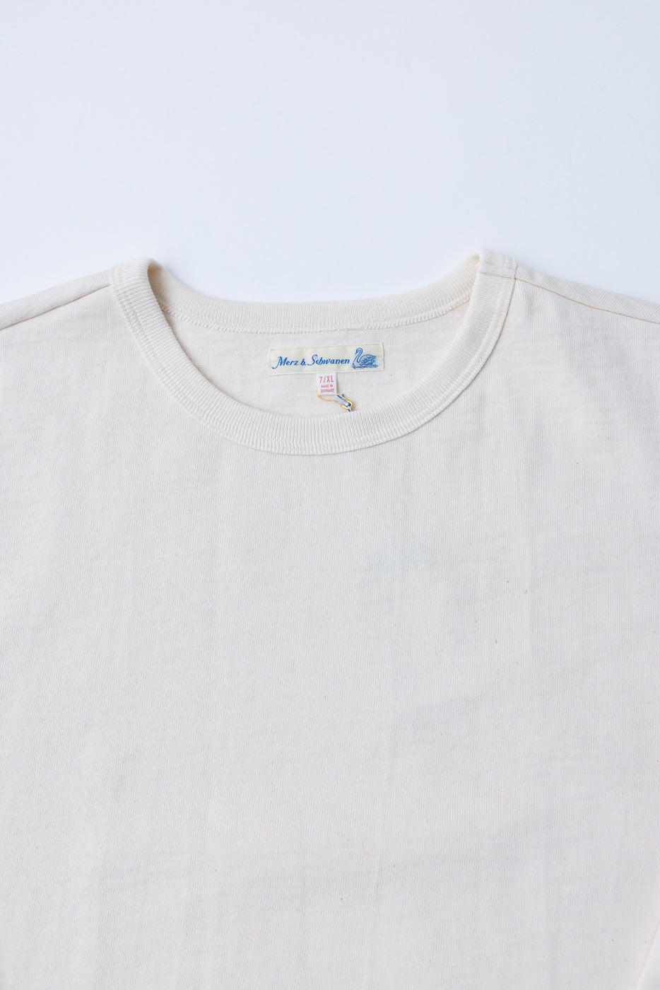 Close-up of the front neckline and tag of a cream white Merz & Schwann t-shirt.