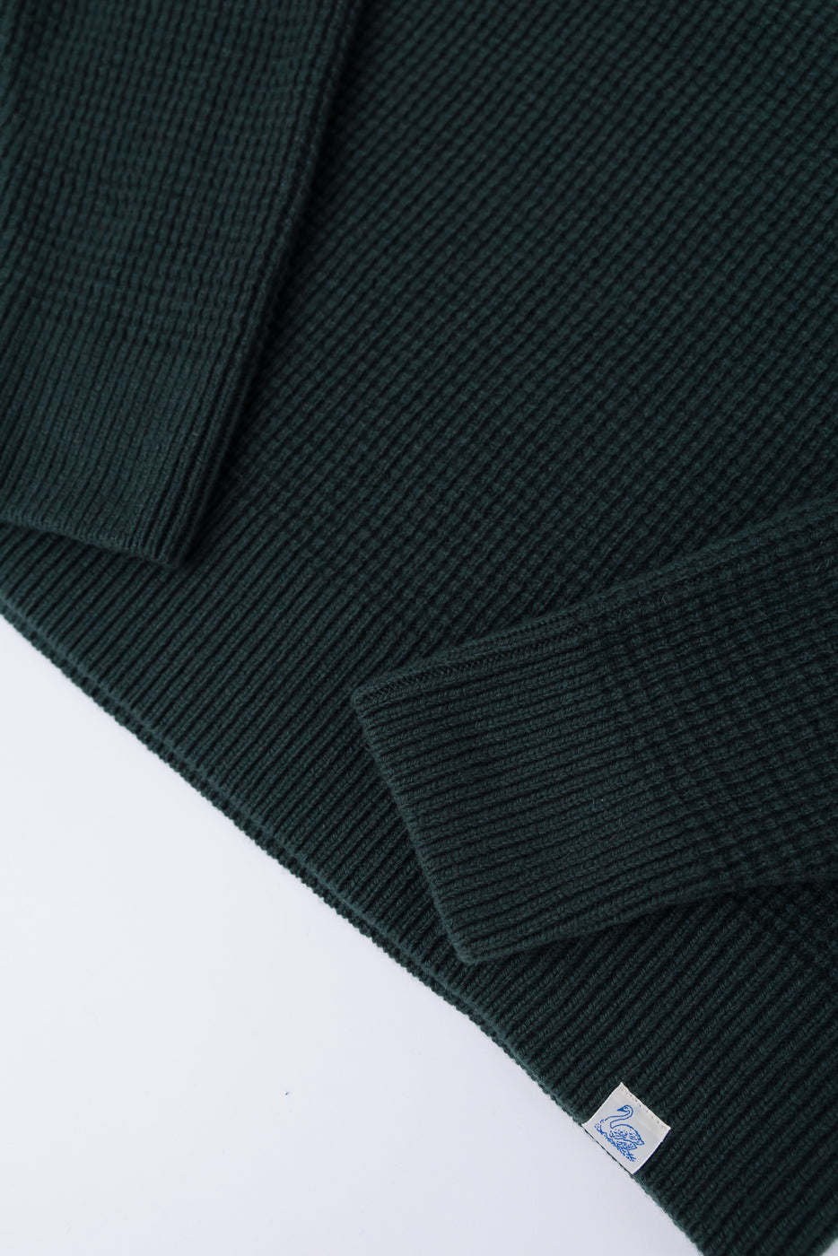 Close-up of bottle green Merz b. Schwanen merino wool cashmere waffle pullover hem and sleeve, front, showing ribbed texture and woven tag.