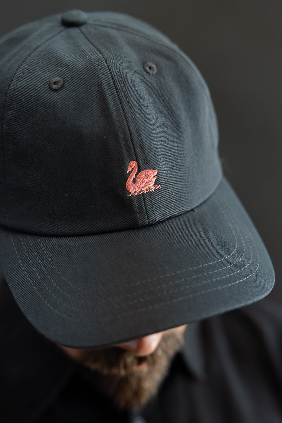Close-up front view of charcoal cotton twill cap with pink swan logo, worn by male model 5'9