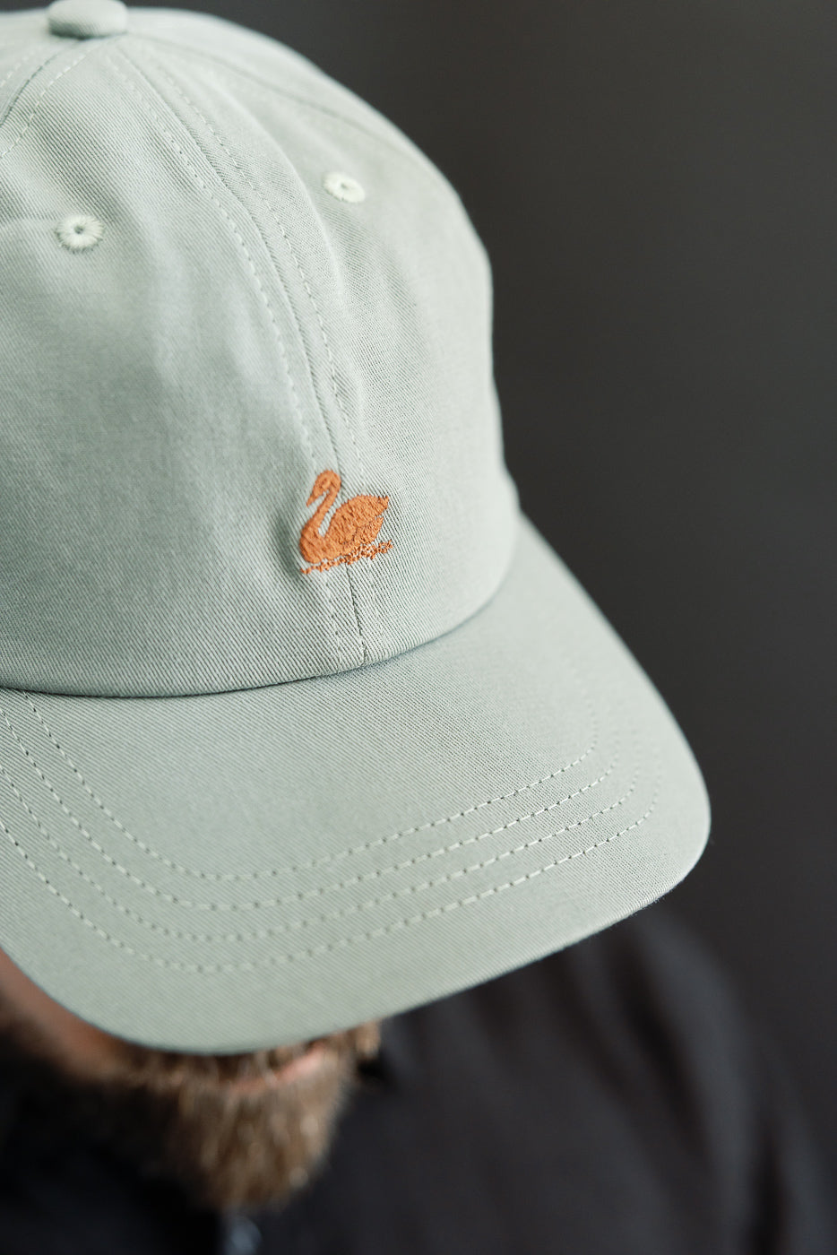 Close-up front view of light army green cotton twill cap with orange swan logo, worn by male model, focusing on embroidery and stitching details.