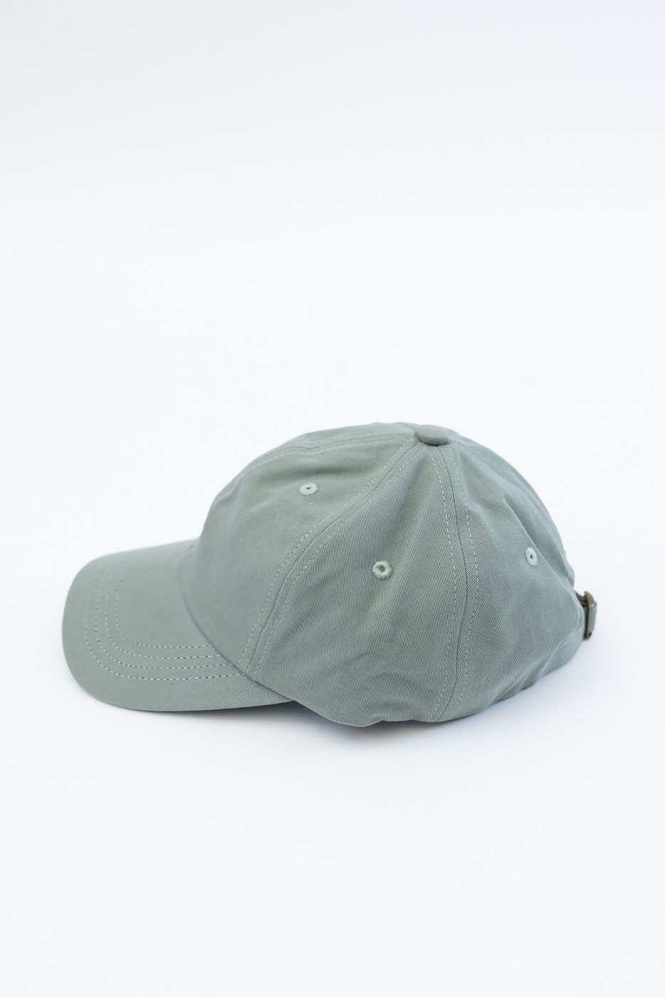 Close-up of light army green cotton twill cap, left side view, showing panel stitching and eyelets.