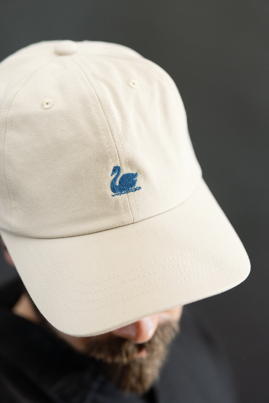 Close-up front view of light khaki cotton twill cap with blue swan logo, worn by male model 5'9