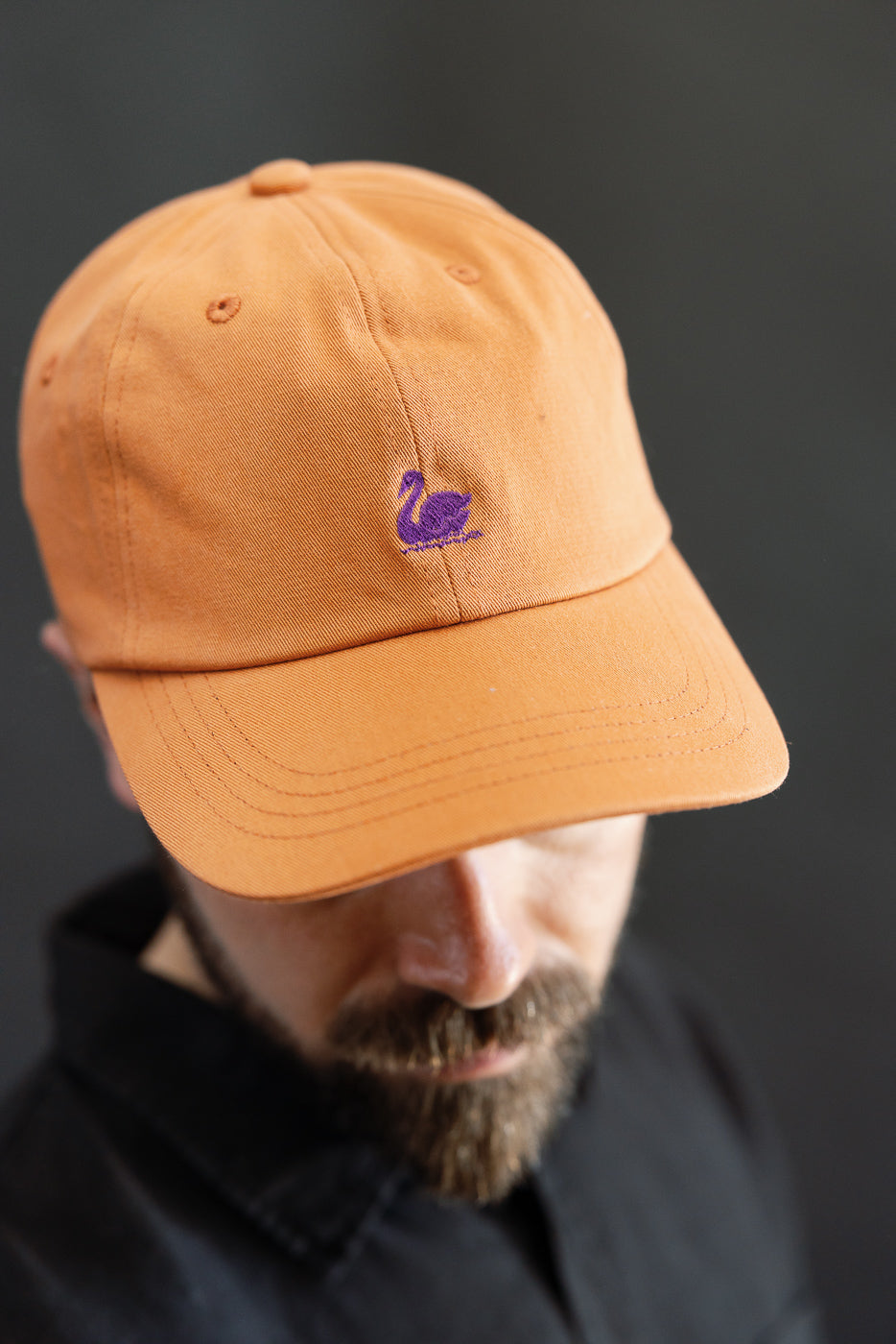 Close-up front view of pumpkin orange cotton twill cap with purple swan logo, worn by male model, 5'9