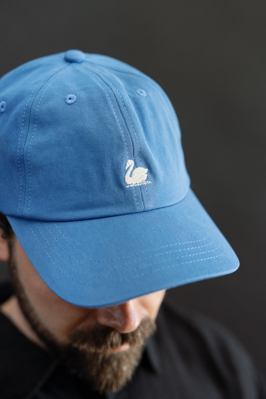 Close-up front view of washed blue cotton twill cap with swan logo, worn by male model 5'9