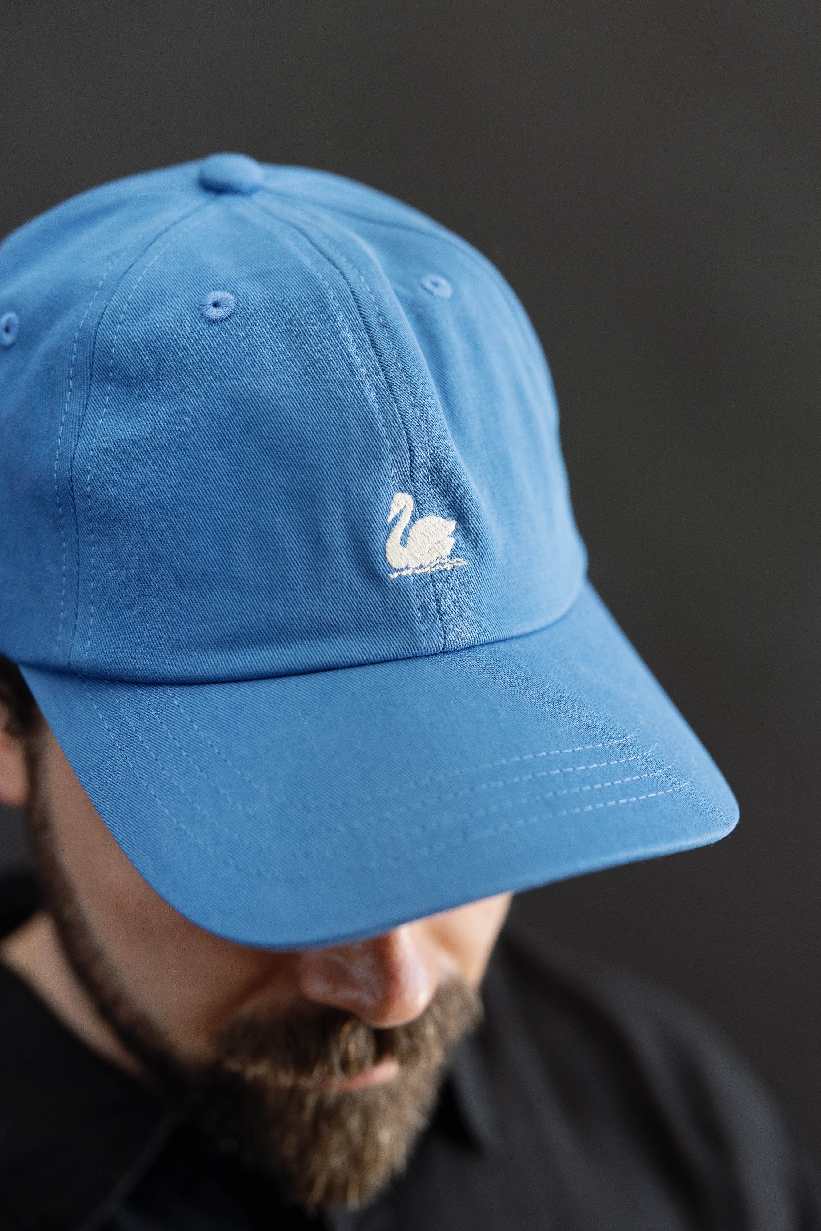 Close-up front view of washed blue cotton twill cap with swan logo on male model, 5'9