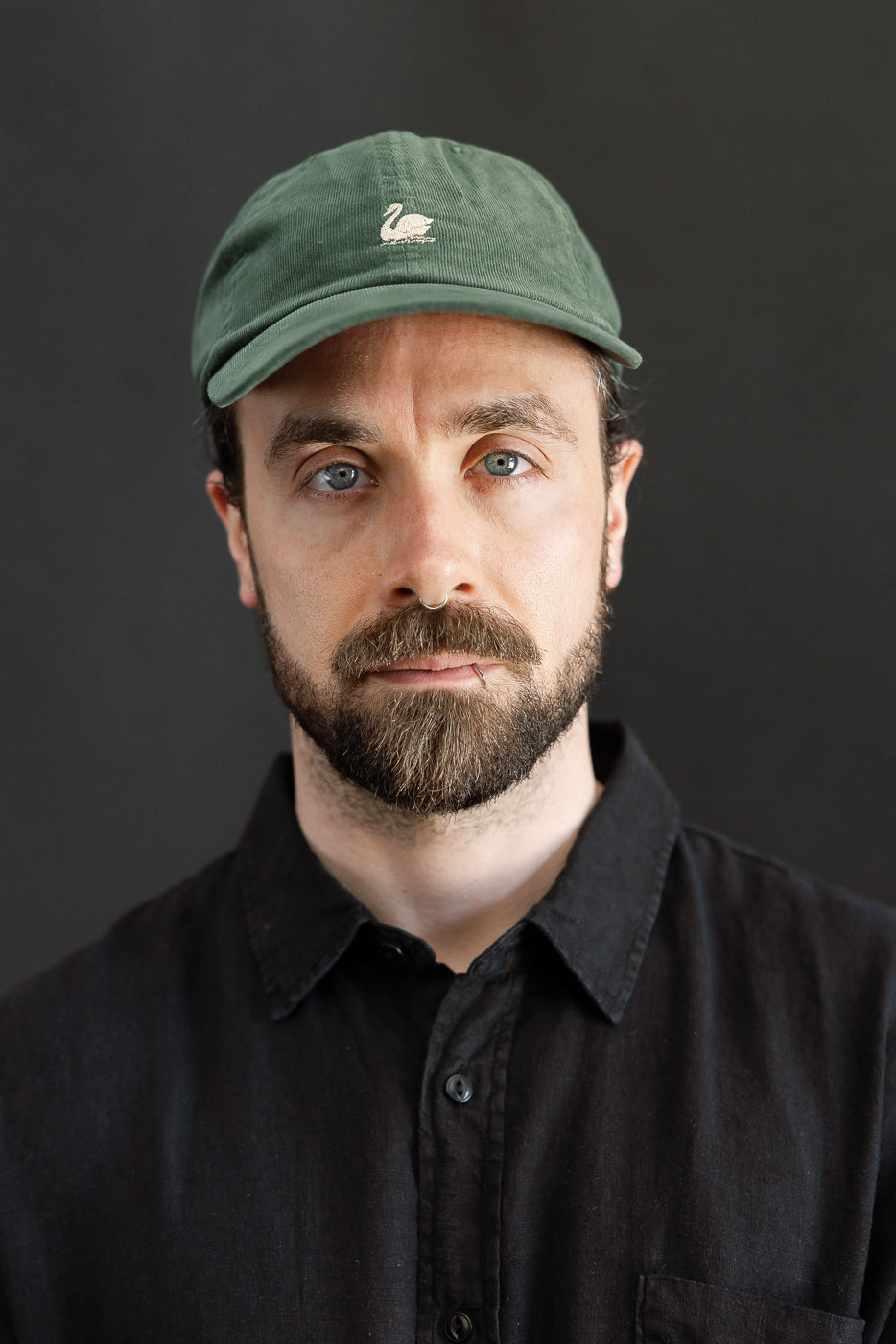 Close-up front view of forest green corduroy cap with swan logo, worn by male model, 5'9