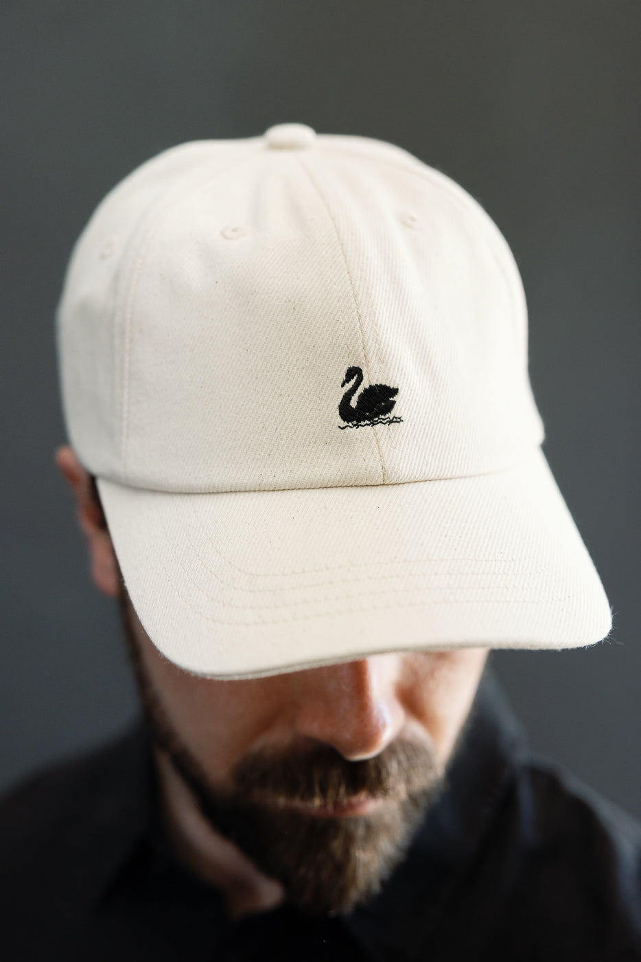 Close-up front view of cream cotton twill cap with black swan logo, worn by male model, 5'9