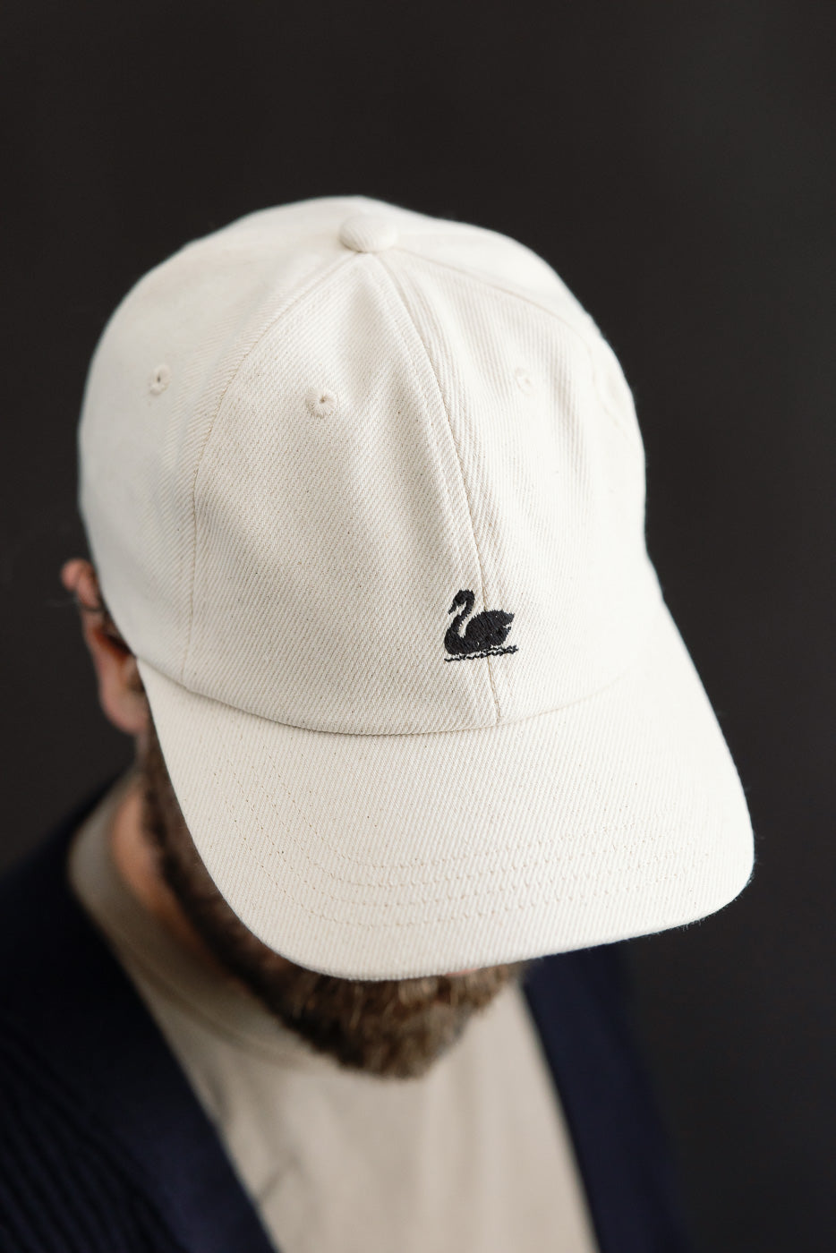Close-up front view of cotton twill cap in natural color with black swan logo, worn by male model, 5'9