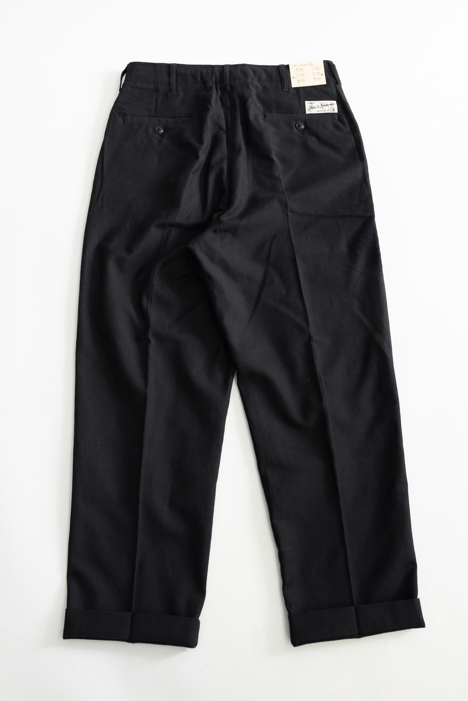 Flat lay of deep black wool cotton worker chino pants, back view, showing cuffed hems, buttoned back pockets, and tag detail.