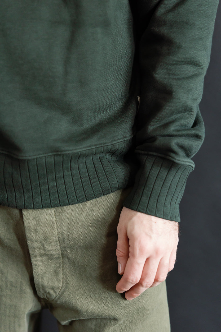 Close-up of ribbed hem and sleeve cuff on heavyweight forest green cotton fleece sweatshirt, male model, front view.