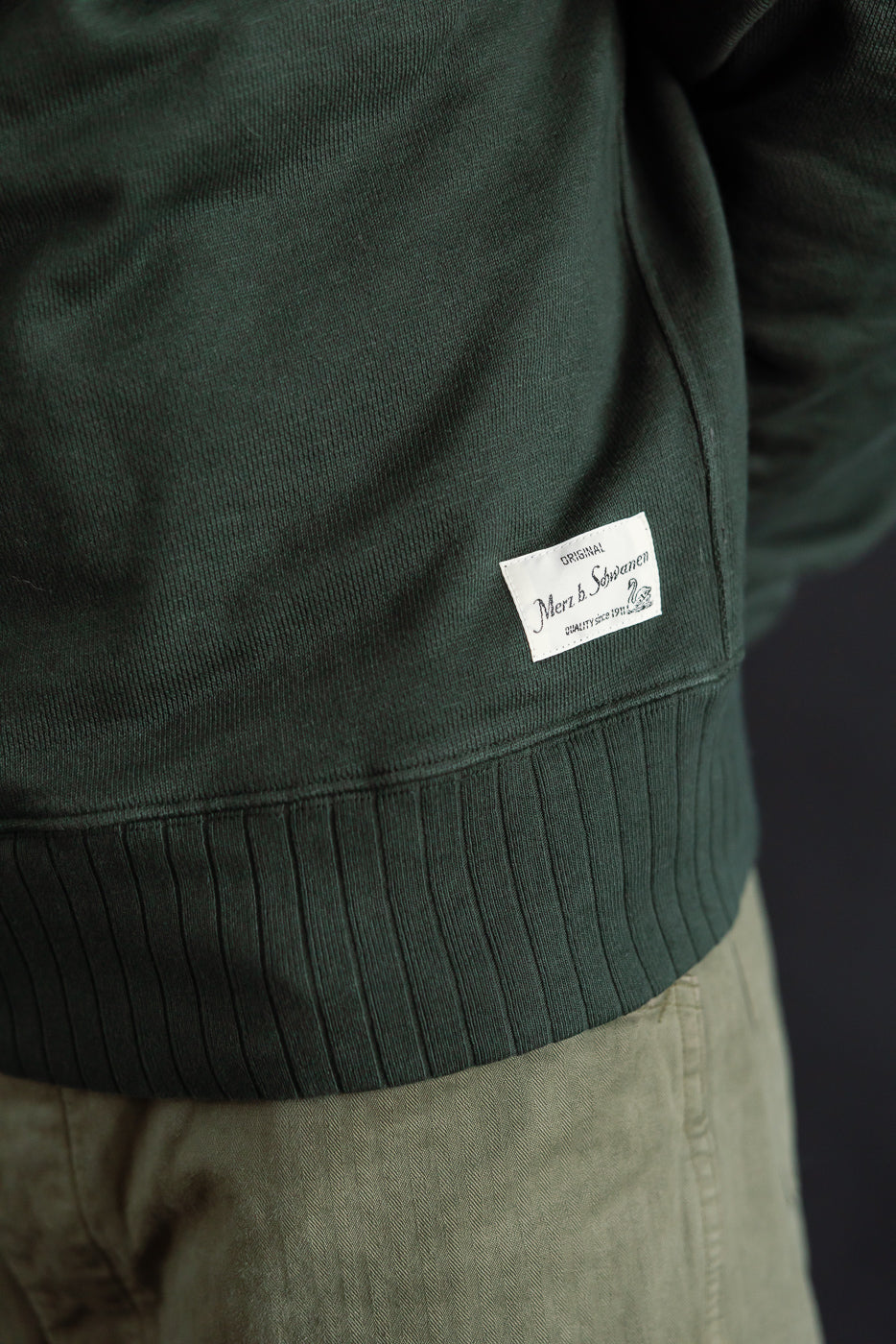 Close-up of men's forest green Merz b. Schwanen heavyweight fleece sweatshirt hem and tag, side view, on male model 5'9